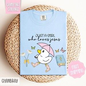 Puede incluir: Camiseta azul claro con un pato de dibujos animados que lleva botas de lluvia y sostiene un paraguas. El texto "Just a girl who loves Jesus" est&aacute; impreso en la camiseta.