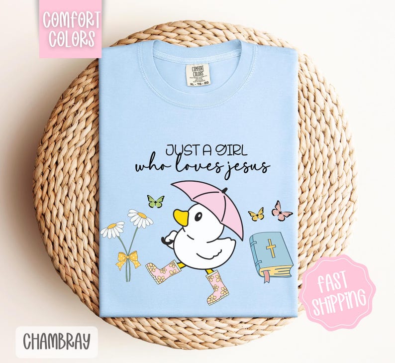 Puede incluir: Camiseta azul claro con un pato de dibujos animados que lleva botas de lluvia y sostiene un paraguas. El texto "Just a girl who loves Jesus" est&aacute; impreso en la camiseta.