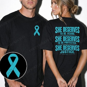 May include: A black t-shirt featuring a teal awareness ribbon on the front chest. The back displays the teal text: SHE DESERVES TO BE HEARD. SHE DESERVES TO BE PROTECTED. SHE DESERVES JUSTICE. PROVERBS 31:8-9. A circular inset shows the teal ribbon.