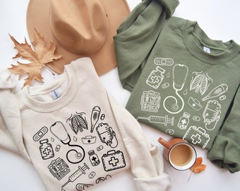 May include: Two cream-colored sweatshirts with a white graphic design of medical symbols, including a stethoscope, a bandage, a thermometer, and a first aid kit. The sweatshirts are laid out on a white surface with a brown hat and a cup of coffee.