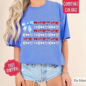 May include: Flo blue t-shirt with a bow-themed design. The design features a large blue bow in the upper left corner, with rows of red and white bows arranged to resemble a flag. The shirt is made of a soft material.