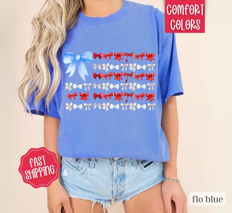 May include: Flo blue t-shirt with a bow-themed design. The design features a large blue bow in the upper left corner, with rows of red and white bows arranged to resemble a flag. The shirt is made of a soft material.