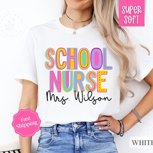 Puede incluir: Camiseta blanca con el texto "SCHOOL NURSE Mrs. Wilson" en letras de colores y contorneadas. La camiseta la lleva puesta una persona con el pelo largo y rubio.