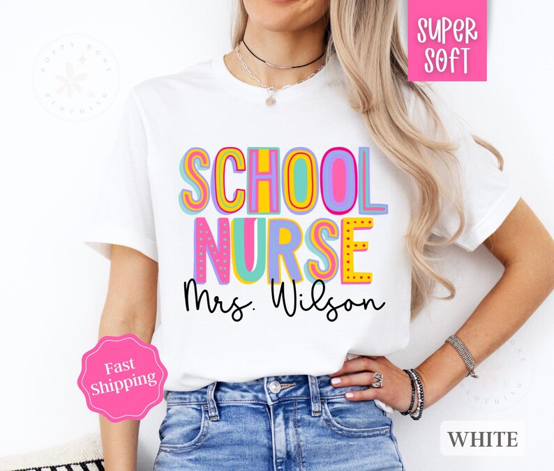 Puede incluir: Camiseta blanca con el texto "SCHOOL NURSE Mrs. Wilson" en letras de colores y contorneadas. La camiseta la lleva puesta una persona con el pelo largo y rubio.