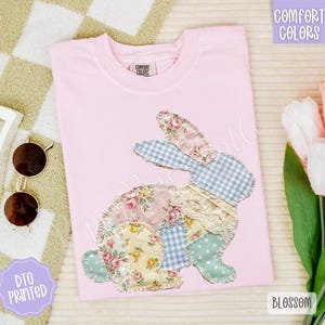 May include: A light pink t-shirt with a patchwork bunny design. The bunny is made of floral, gingham, and lace fabrics. The shirt has the words "Comfort Colors" printed on it. Sunglasses and a hair clip are on the left.