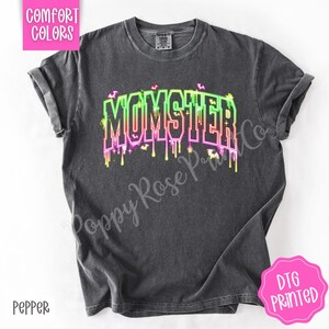 May include: A dark gray t-shirt with the word "MOMSTER" in a neon green, pink, and yellow dripping font. The shirt has a crew neck and short sleeves. The text "Comfort Colors" is in a pink box in the upper left corner, and "DTG PRINTED" is in a pink circle in the lower right corner.