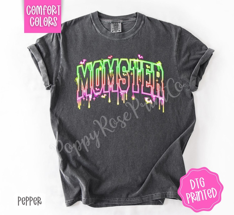 May include: A dark gray t-shirt with the word "MOMSTER" in a neon green, pink, and yellow dripping font. The shirt has a crew neck and short sleeves. The text "Comfort Colors" is in a pink box in the upper left corner, and "DTG PRINTED" is in a pink circle in the lower right corner.