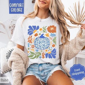 Op de afbeelding: Wit T-shirt met een blauw en wit pompoenontwerp, omringd door kleurrijke bloemen en bladeren. De tekst "Comfort Colors" is zichtbaar in de linkerbovenhoek van de afbeelding.