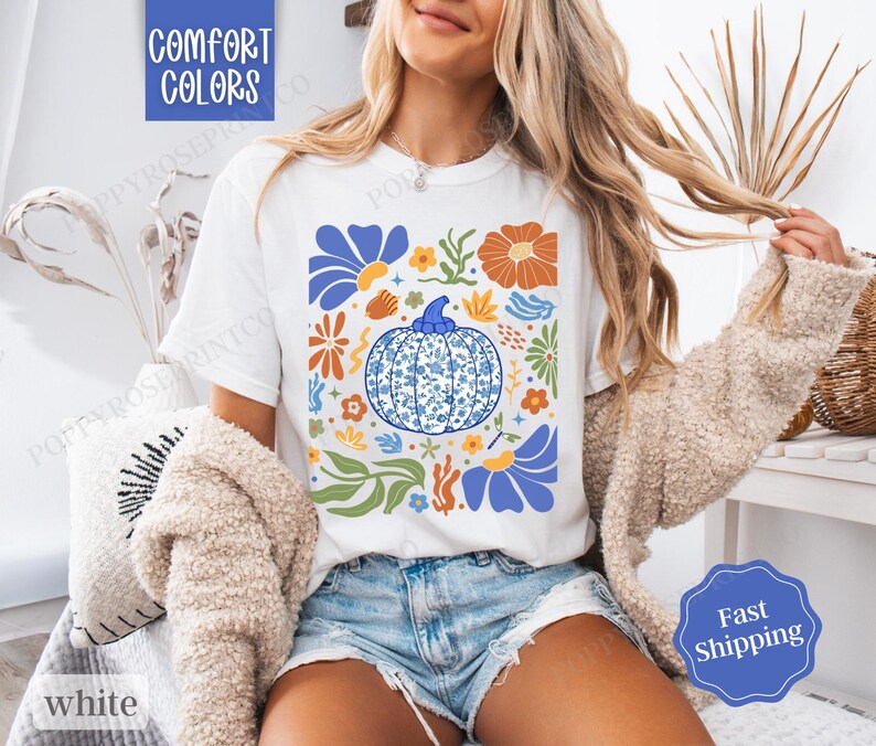 Op de afbeelding: Wit T-shirt met een blauw en wit pompoenontwerp, omringd door kleurrijke bloemen en bladeren. De tekst "Comfort Colors" is zichtbaar in de linkerbovenhoek van de afbeelding.