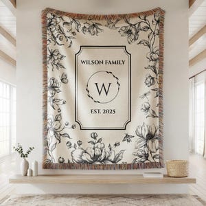 May include: Cream-colored woven throw blanket with a floral design and the text "WILSON FAMILY", a "W" inside a circle, and "EST. 2025". The blanket has a fringed edge and is decorated with black and white flowers, butterflies, and bees.
