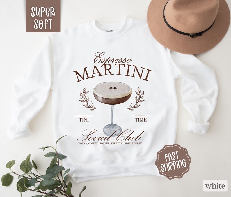 May include: White sweatshirt with brown text that reads "Espresso Martini" and a graphic of a martini glass with coffee beans. The text below the graphic reads "Tini Time Social Club Vodka, Coffee Liqueur, Espresso, Simple Syrup".