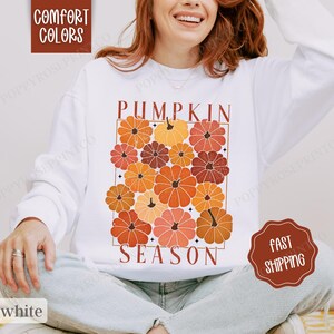 May include: White sweatshirt with the text "Pumpkin Season" in a retro font. The text is surrounded by a graphic of orange, red, and brown pumpkins.