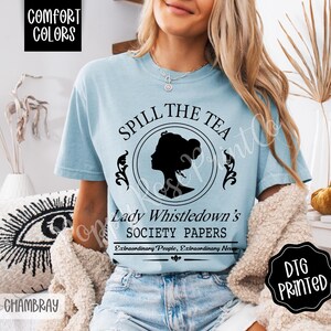 Pu&ograve; includere: T-shirt azzurra con la scritta "SPILL THE TEA" e "Lady Whistledown's SOCIETY PAPERS". La maglietta presenta una silhouette nera del profilo di una donna all'interno di un disegno circolare. La maglietta &egrave; in tessuto chambray.