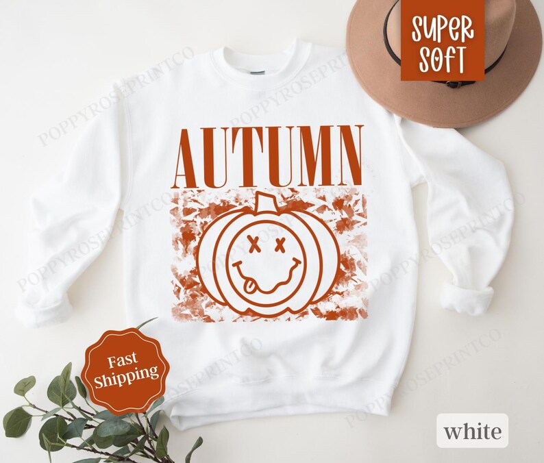 May include: White sweatshirt with a brown graphic of a smiling pumpkin with the word "AUTUMN" above it.
