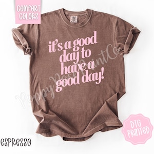 Puede incluir: Camiseta color espresso con la frase rosa "it's a good day to have a good day!". La camiseta tiene mangas cortas y cuello redondo. Las palabras "COMFORT COLORS" y "DTG PRINTED" también son visibles.