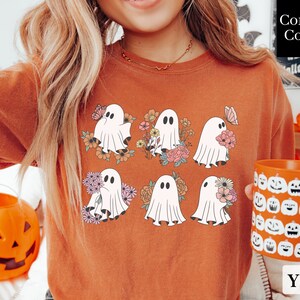May include: A rust-colored t-shirt with a graphic of six white ghosts holding flowers. The ghosts are surrounded by floral designs in shades of pink, yellow, and green. The text "Comfort Colors" is visible in the upper right corner of the image.