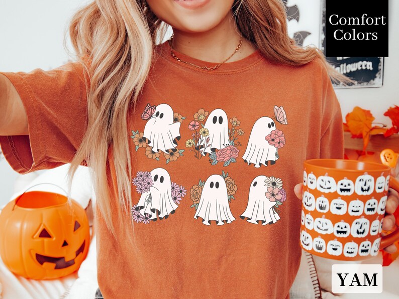 May include: A rust-colored t-shirt with a graphic of six white ghosts holding flowers. The ghosts are surrounded by floral designs in shades of pink, yellow, and green. The text "Comfort Colors" is visible in the upper right corner of the image.