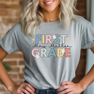 Op de afbeelding: Een lichtgrijs T-shirt met de tekst "FIRST Mrs. Miller GRADE" in kleurrijke blokletters met bloemen en glitteraccenten.
