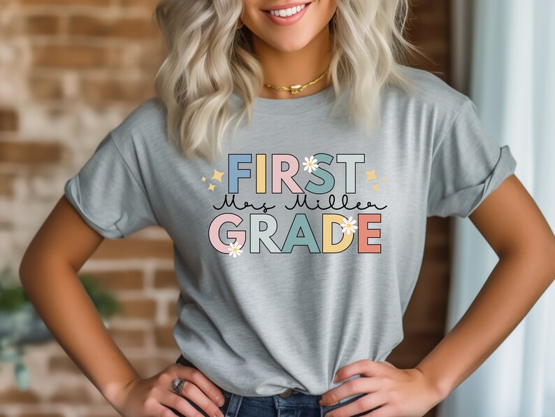 Op de afbeelding: Een lichtgrijs T-shirt met de tekst "FIRST Mrs. Miller GRADE" in kleurrijke blokletters met bloemen en glitteraccenten.