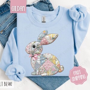 May include: A light blue Gildan sweatshirt with a patchwork bunny design. The bunny is made up of different colored fabric squares and has a pink bow around its neck.