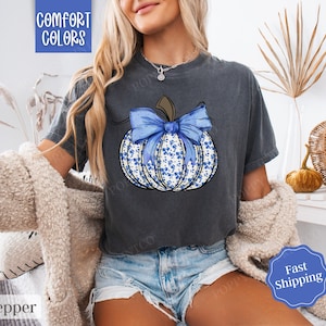 May include: A dark grey t-shirt featuring a blue and white floral pumpkin with a blue bow. The text "Comfort Colors" is in the top left corner of the shirt.
