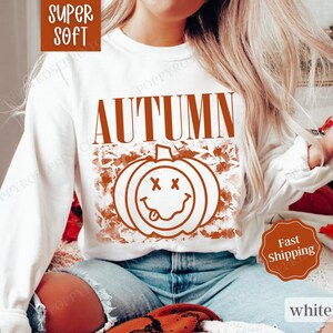 May include: White long-sleeved sweatshirt with the word "AUTUMN" in orange text and a smiling pumpkin graphic with an orange background.