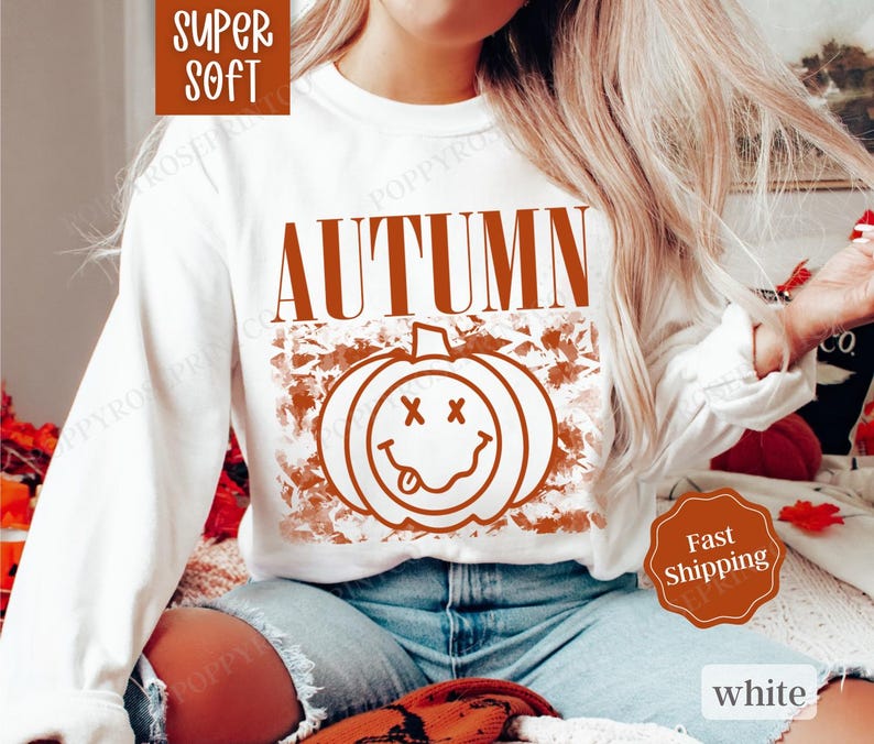 May include: White long-sleeved sweatshirt with the word "AUTUMN" in orange text and a smiling pumpkin graphic with an orange background.