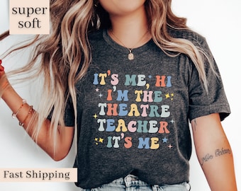 May include: A dark gray t-shirt with the text "IT'S ME, HI I'M THE THEATRE TEACHER IT'S ME" in colorful retro-style lettering.