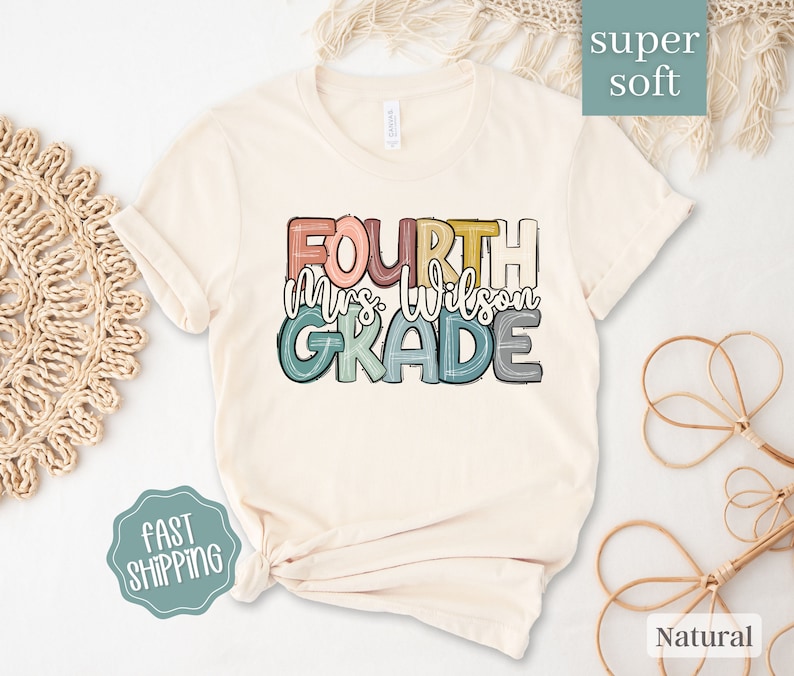 May include: A beige t-shirt with the text "FOURTH mrs. wilson GRADE" in colorful, stylized lettering. The shirt is made of a soft, comfortable fabric.