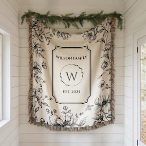May include: Cream-colored throw blanket with a floral design and fringe. The center features a framed design with "WILSON FAMILY", a "W" in a circle, and "EST. 2025". The blanket is decorated with black and white floral illustrations.