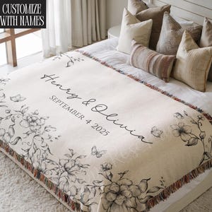 May include: Cream-colored throw blanket with floral and butterfly illustrations. The blanket features the names "Henry & Olivia" and the date "SEPTEMBER 4 2025" in black script. The text "CUSTOMIZE WITH NAMES" is in the upper left corner.