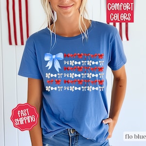 May include: Flo blue t-shirt with a bow and ribbon design in red, white, and blue. The shirt has the words "Comfort Colors" and "flo blue" printed on it. The shirt also has a "Fast Shipping" sticker.