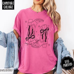 May include: A pink t-shirt with a black graphic of two cowboy boots and the text "Long Live Cowgirls". The shirt has short sleeves and a relaxed fit. The words "Comfort Colors" and "DTG Printed" are also visible.