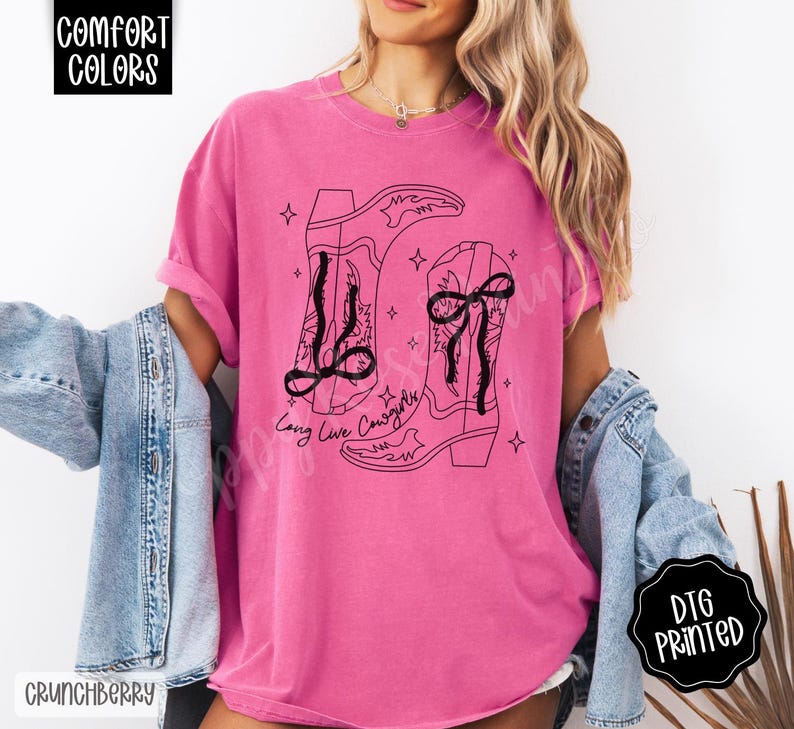 May include: A pink t-shirt with a black graphic of two cowboy boots and the text "Long Live Cowgirls". The shirt has short sleeves and a relaxed fit. The words "Comfort Colors" and "DTG Printed" are also visible.