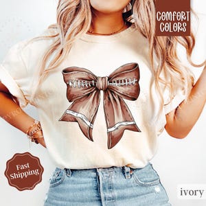 May include: A white t-shirt with a brown bow design featuring football stitching. The bow is centered on the shirt and has a cream-colored background. The text "COMFORT COLORS" is in a brown rectangle at the top of the image. The text "ivory" is in a brown rectangle at the bottom of the image.
