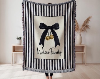 May include: A black and white striped throw blanket with a central design featuring a black bow, two gold bells, and the text "Wilson Family". The blanket has a fringed edge and is displayed indoors.