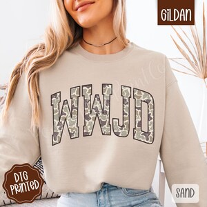 May include: A sand-colored sweatshirt with the text "WWJD" in a camouflage pattern. The sweatshirt is a crew neck style and has long sleeves. The image also includes the Gildan logo and the word "Sand".
