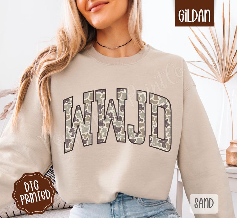 May include: A sand-colored sweatshirt with the text "WWJD" in a camouflage pattern. The sweatshirt is a crew neck style and has long sleeves. The image also includes the Gildan logo and the word "Sand".