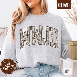 May include: A light gray sweatshirt with the letters "WWJD" in a camouflage pattern across the chest. The sweatshirt is a Gildan brand and has a DTG printed logo.
