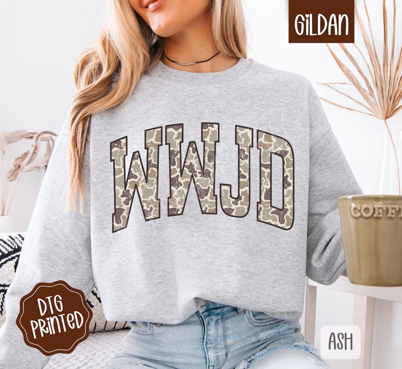 May include: A light gray sweatshirt with the letters "WWJD" in a camouflage pattern across the chest. The sweatshirt is a Gildan brand and has a DTG printed logo.