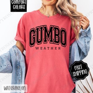 May include: A red t-shirt with the text "GUMBO WEATHER" in black block letters. The shirt is made of a soft, comfortable fabric.