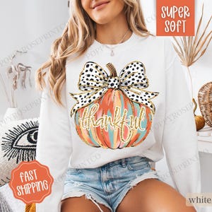 May include: White sweatshirt featuring a graphic of a pumpkin with a black and white polka dot bow. The pumpkin is painted in shades of orange, pink, and blue. The word "thankful" is written in gold glitter on the pumpkin.