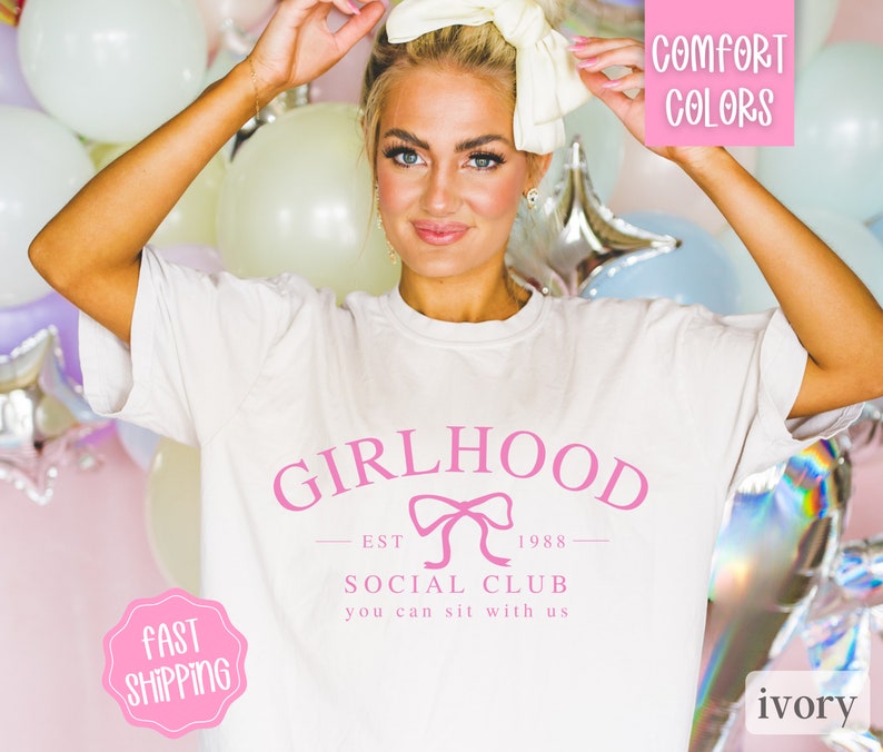 May include: White t-shirt with a pink graphic that says "Girlhood Social Club Est. 1988 You can sit with us".