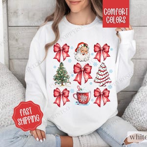May include: White sweatshirt with a Christmas theme featuring red bows, a Santa Claus face, a Christmas tree, and a cup of hot chocolate. The text "Comfort Colors" is on a red tag attached to the sweatshirt.