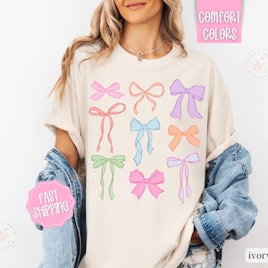 May include: Ivory-colored t-shirt featuring a collection of colorful bow illustrations. The bows are in various pastel shades, including pink, blue, and green. The shirt has the words "Comfort Colors" and "Fast Shipping" on it.