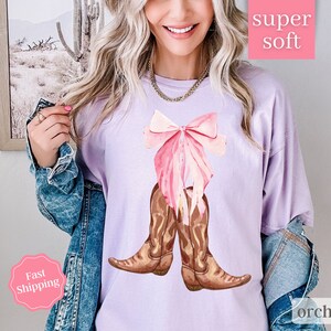 K&ouml;nnte beinhalten: Ein lavendelfarbenes T-Shirt mit einem rosa Schleifen- und braunen Cowboy-Stiefel-Motiv. Der Text "super soft" befindet sich in einem rosa Kasten &uuml;ber dem Motiv und der Text "orchid" in einem rosa Kasten unter dem Motiv.