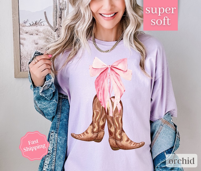 K&ouml;nnte beinhalten: Ein lavendelfarbenes T-Shirt mit einem rosa Schleifen- und braunen Cowboy-Stiefel-Motiv. Der Text "super soft" befindet sich in einem rosa Kasten &uuml;ber dem Motiv und der Text "orchid" in einem rosa Kasten unter dem Motiv.