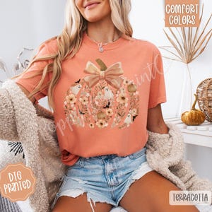 Pu&ograve; includere: T-shirt color terracotta con un design a forma di zucca piena di fiori, fantasmi e un fiocco. La maglietta ha le parole "Comfort Colors" e "DTG Printed" stampate.