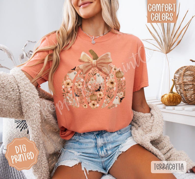 Pu&ograve; includere: T-shirt color terracotta con un design a forma di zucca piena di fiori, fantasmi e un fiocco. La maglietta ha le parole "Comfort Colors" e "DTG Printed" stampate.