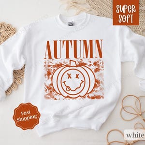 May include: White sweatshirt with the text "AUTUMN" in orange and a graphic of a smiling pumpkin with a tongue sticking out.